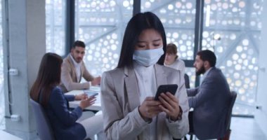 Asyalı genç bir kadın, sağlık maskeli beyaz yakalı bir ofis çalışanı ofiste duruyor ve akıllı telefondan mesaj yolluyor. Kadın şirket çalışanı telefonda dinleme ve kaydırma yapıyor. İş toplantısı geçmişi.
