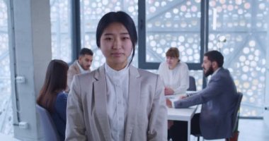 Ofiste durmuş gülümseyen kameraya bakan Asyalı genç bir ofis çalışanının portresi. Arka planda iş toplantısı. Şirketin kadın çalışanı mutlu bir şekilde gülümsüyor. Kız stajyer konsepti.