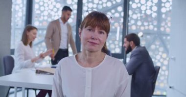 Kabinede durup kameraya bakan orta yaşlı beyaz kadın ofis çalışanı portresi. Arka planda iş toplantısı. Kafasında gözlük olan firmanın kadın çalışanı. Beyaz yaka konsepti.