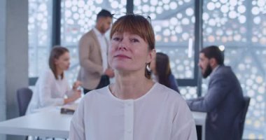 Kabinede durup kameraya gülümseyen orta yaşlı beyaz kadın ofis çalışanı portresi. Arka planda iş toplantısı. Firmanın kadın çalışanı gözlük takıyor ve gülüyor..