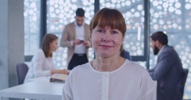 Kameraya bakıp gülümseyen Kafkas kadın ofis çalışanı portresi. Arka planda iş toplantısı. Gülümseyen orta yaşlı kadın şirket çalışanı. Beyaz yakalı personel konsepti.
