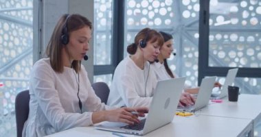 Kulaklıklı genç beyaz kadın diz üstü bilgisayarda yazı yazıyor ve çağrı merkezinde müşteriyle konuşuyor. Müşteri destek bürosunun kadın operatörleri. Kız çevrimiçi teknik sorunu çözmeye yardım ediyor.