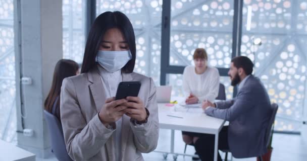 オフィスに立ってスマートフォンにメッセージを送る女性オフィスワーカー 会社のトレーナーがタップして電話をスクロールします 医療マスクでアジアの若い女性の肖像画 ビジネスミーティングの背景 — ストック動画
