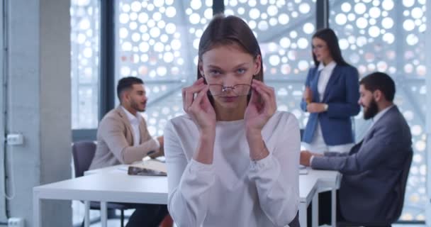 キャビネットに立ってカメラを見ている若い白人女性オフィスワーカーの肖像画 バックグラウンドでのビジネスミーティング メガネをかけている会社の女性社員 女子トレーニングコンセプト — ストック動画