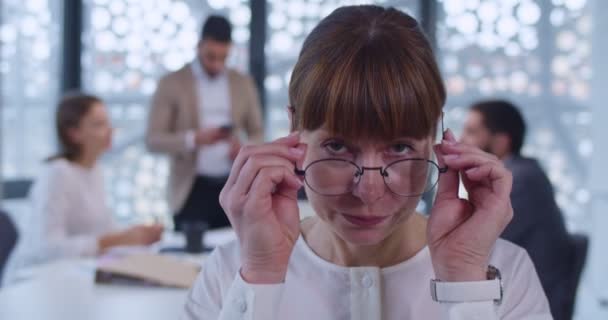 接近中年白种人女性上班族的镜头看着相机 关于背景的商务会议 工作场所戴眼镜的公司高级女员工 — 图库视频影像