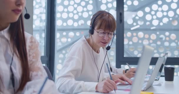 在服务台笔记本电脑呼叫中心工作的耳机中的白人妇女 技术支助机构的女性同事与客户交谈 帮助解决问题和销售 — 图库视频影像