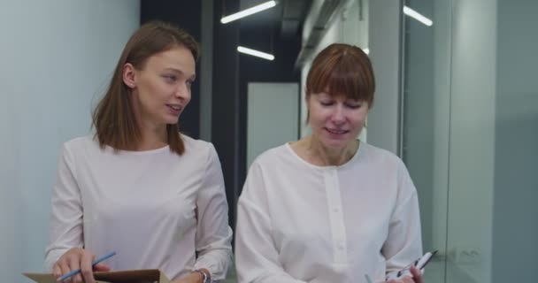 两个白人妇女 年轻的和中年的白领同事 坚定地走过走廊 谈论着工作 同事中的女性同事在办公室里聊天和谈论工作 — 图库视频影像