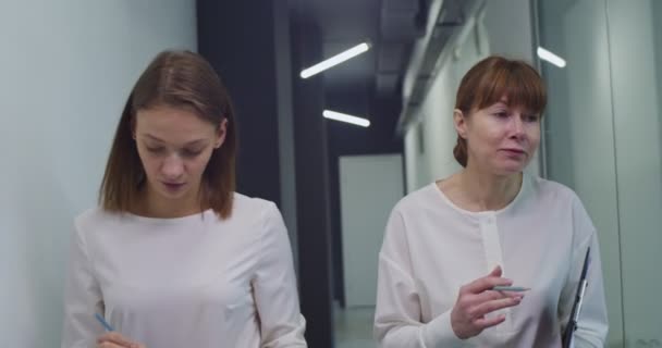 两个白人妇女 年轻的和中年的白领同事 在走廊上与人同行 谈论工作 与同事聊天和上班的女性 — 图库视频影像