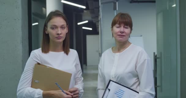 两个白人妇女的画像 年轻的和中年的白领同事站在公司走廊里 手里拿着文件夹 看着相机 在职的女性同事 — 图库视频影像