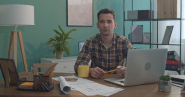 Beyaz, genç, yakışıklı bir ofis çalışanı dizüstü bilgisayarla masada oturuyor ve kameraya bakıyor. Portre çekimi.