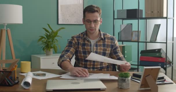 ガラス張りの若い白人男性デザイナーがアパートのインテリアプランを描き 写真付きのシートに鉛筆を描く — ストック動画