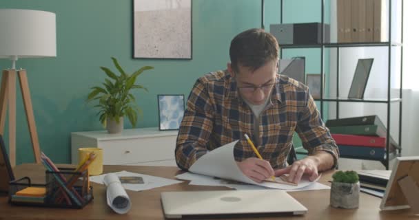 年轻英俊的白人男子坐在房间的办公桌前 用铅笔和三角形画出图画或建筑草图 — 图库视频影像