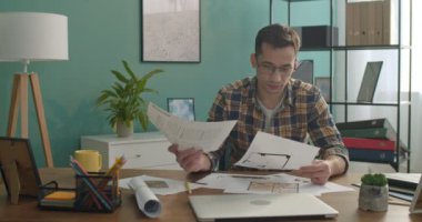 Gözlüklü beyaz adam mimar veya tasarımcı çalışma masasındaki taslaklara, grafiklere, çizimlere ve planlara bakıyor.