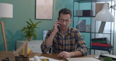 Gözlüklü genç beyaz adam ofis odasında ya da evde masada oturuyor ve cep telefonuyla konuşuyor..