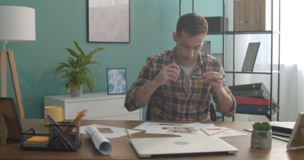 年轻的白人建筑师或设计师戴着眼镜 看着工作台旁的摄像机 桌子上有漂亮房间里的草稿 — 图库视频影像