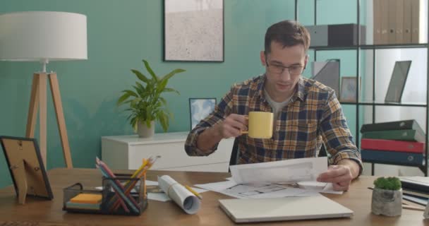 白种人年轻英俊的建筑师或戴眼镜的设计师 在办公桌前与草图和图形一起工作 喝热茶或咖啡 — 图库视频影像