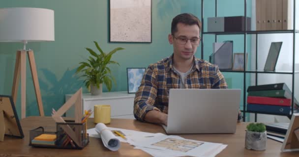 照片上的这位年轻的白种人英俊男子戴着眼镜 在办公室或家里的笔记本电脑前工作 对着摄像机微笑 — 图库视频影像