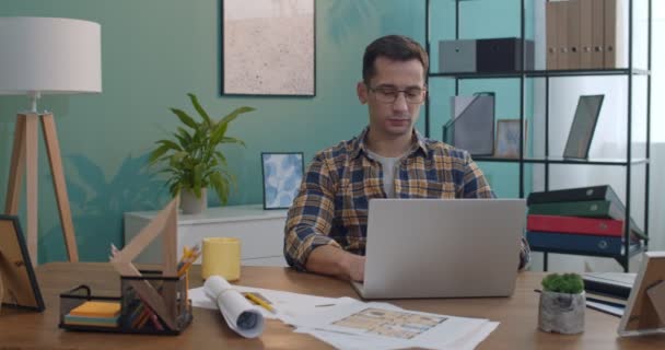 年轻的白种人戴眼镜 一边在办公室桌上的笔记本电脑前工作 一边在家里喝热茶或咖啡 — 图库视频影像
