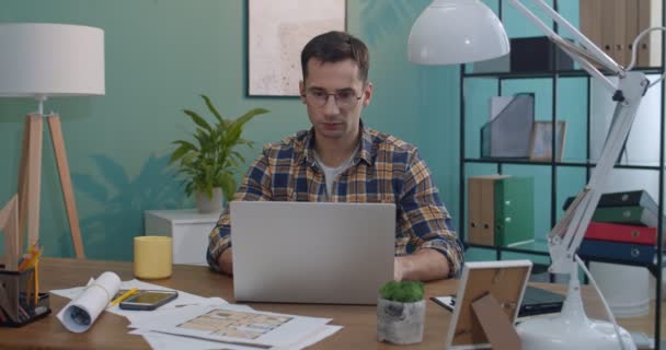 白种人年轻英俊的男人戴着眼镜 在办公室或家里的笔记本电脑前工作 然后拿着智能手机在上面看书或发短信 — 图库视频影像