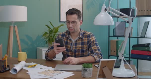 白种人年轻英俊的男人戴着眼镜 坐在办公室或家里的桌子旁 用智能手机敲敲或发短信 — 图库视频影像