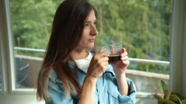 Neşeli, beyaz, güzel bir kadın sabah elinde kahve ya da çayla pencerenin yanında durup gülümsüyor. Boş zaman konsepti