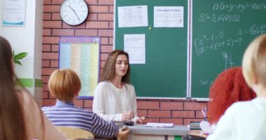 Genç, beyaz, güzel bayan öğretmen küçük öğrencilere matematiği kara tahtada açıklıyor. Masalarda oturup matematik dersi dinleyen çocukların arkası. Arkadan bak. Öğretim ve öğrenme.