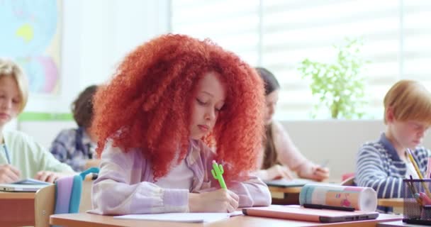 可爱的白种人小女孩 一头卷曲的红头发坐在课桌前 听老师讲课 用笔在抄本上写字 在学校学习的小女生 学生学习概念 — 图库视频影像