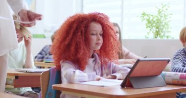 Sınıftaki küçük şirin beyaz kız öğrencinin tablet bilgisayarlı portresi. Öğretmen sınav kâğıdına görev veriyor, kız yazmaya başlıyor. Cihazla konsept öğrenme. Okul hayatı.