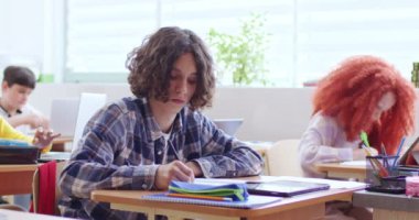 Öğretmenleri dinleyen ve ilkokulda ders sırasında not defterine yazma alıştırmaları yapan ciddi anaokulu öğrencileri. Dijital tablet kullanan kıvırcık saçlı düşünceli bir çocuk..