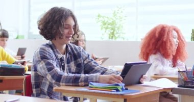 Kıvırcık saçlı, elinde tabletle sınıftaki masasında oturan ve ders sırasında gülümseyen genç bir erkek. Tanımlanamayan öğretmen yürüyor ve öğrenciyi azarlıyor..