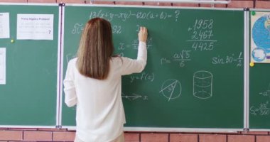 Yeşil karatahtanın yanında duran ve sınıfta geometrik formül yazan kadın öğretmen. Matematik kurallarını çözen ve açıklayan okul öğretmeni.