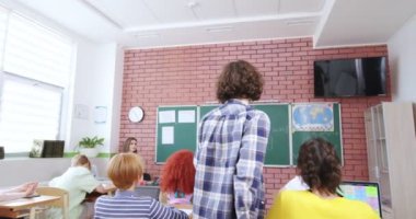 Sınıfın masasından kalkıp tahtaya giden beyaz bir gencin arkası. Bayan öğretmen matematik denklemleri için okul çocuğunu arıyor. Sınıfta çocuklarla arka plan görüntüsü.