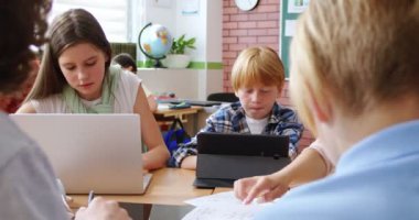 Gülümseyen öğretmen sınıfta masada otururken okul çağındaki çocuklara yeni materyalleri açıklıyor. Ders sırasında dizüstü bilgisayar ve tablet kullanan bir grup öğrenci.