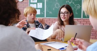 Beyaz kadın öğretmen ve bir grup öğrenci dizüstü bilgisayarları ve tabletleriyle masada oturuyorlar. Sınıftaki kız ve oğlanlara kimyayı açıklayan mutlu öğretmen..