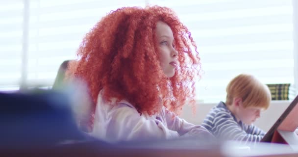 在明亮的教室里 一个红头发的女学生坐在桌子旁 手里拿着平板电脑 无法辨认的女教师走路和分发有考试任务的工作单 — 图库视频影像