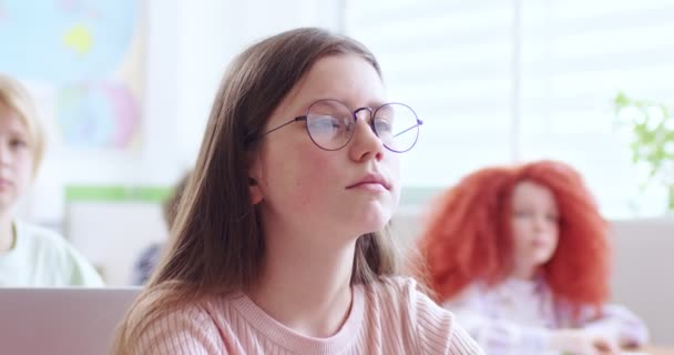 一个漂亮的白种人少女的画像 她戴着眼镜 坐在教室的桌子旁边 看着镜头在上课 在孩子们中间室内 小女孩在学习学校的概念 靠近点 — 图库视频影像