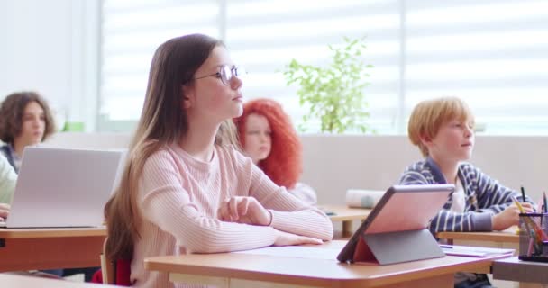 用长长的棕色头发高举着眼镜 等待轮到她回答棘手的问题的可爱女生 孩子们坐在课桌前 手里拿着笔记本电脑和平板电脑 — 图库视频影像