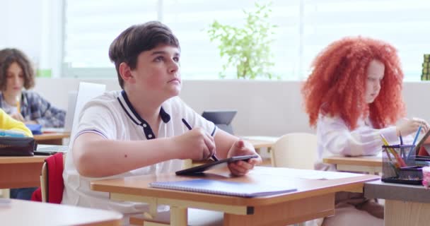 10代の白人の少年が教室の机に座って授業中の子供たちの間で 学習中にタブレットデバイスを使用しています コンピュータの画面上のスマート少年のテキストと学校で黒板を見て — ストック動画