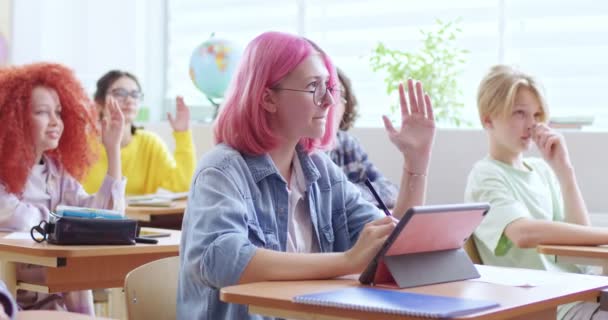 教室のデジタルタブレットで机に座っている眼鏡にピンクの髪のきれいな女の子の肖像画 教師の質問に答えるために手を上げるスマート女性学生 — ストック動画