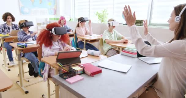 Attractive female teacher in headphones instructing pupils how to swipe on virtual screen in VR headsets. Caucasian children using innovative gadgets for studying at modern school.