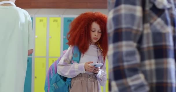 红头发女孩在学校走廊里使用现代智能手机 而其他学生在她身边走来走去 带着背包的女学生在课间休息时独自站着 看着相机 — 图库视频影像