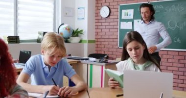 Hevesli beyaz bir öğretmen masasında oturan erkek ve bayan öğrencilere dijital tablet gösteriyor. Modern okulda farklı aletlerin yardımıyla yeni şeyler öğrenen zeki çocuklar..