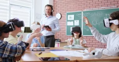 VR kulaklıklı bir grup genç öğrenci masada otururken dijital tabletli erkek öğretmenler eğitim programını açıklıyorlar. Okulda etkileşimli ders sırasında modern teknolojiler.