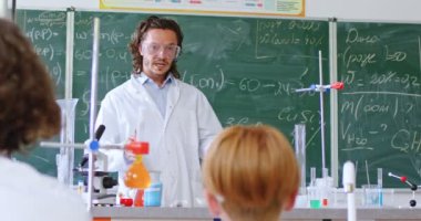 Gözlüklü beyaz erkek öğretmen sınıfta formüllerle karatahtanın başında duruyor, dersleri anlatıyor ve öğrencilere tüplerde kimyasallar gösteriyor. Sınıfta bir deney. İlginç bir çalışma..