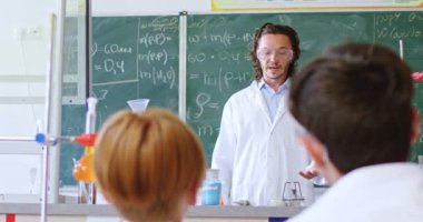 Güvenlik gözlüğü ve laboratuvar önlüğü takmış beyaz bir öğretmen ders sırasında öğrencilere kimyasal reaksiyonlar göstermek için test tüplerinde sıvı karıştırıyor. İlgiyle öğrenmek için bilimsel deneyler.
