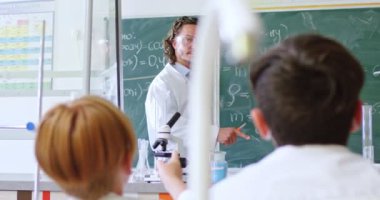 Gözlüklü beyaz erkek öğretmen tahtadaki kimyasal formülleri açıklıyor. Okul dersi konsepti. Genç öğrencilere kimyayı açıklayan adam. Masadaki çocukların arkası. Sınıfın arka görünümü