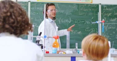 Koruyucu gözlüklü ve beyaz önlüklü bir erkek öğretmen. Tahtanın yanında durup kimyasal formüller yazıyor ve çocuklarla konuşuyor. Kimya dersi sırasında masada oturan öğrenciler..