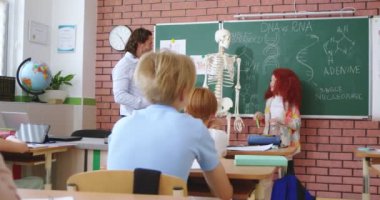 Kızıl saçlı liseli kız, erkek öğretmene sorularını cevaplarken iskelette insan parçaları gösteriyor. Modern okulda biyoloji dersi. Faiziyle eğitim.