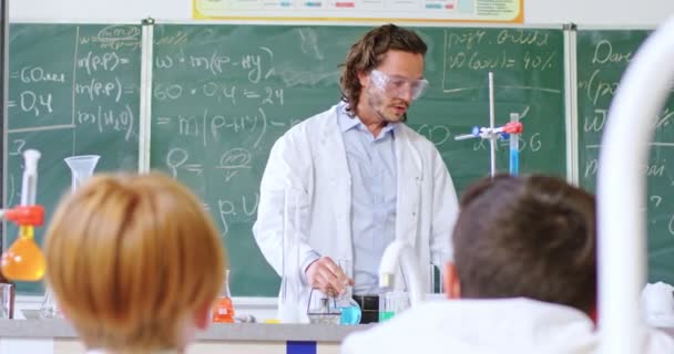 学校化学课 身穿护目镜的白人男教师站在黑板前 课堂上带着配方 并向学生展示管子中的化学物质 实验证明 有趣的研究 — 图库视频影像