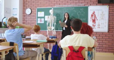 Anaokulu öğrencilerine anatomi dersi vermek için iskelet modeli kullanan pozitif bir kadın. Çocuklar ders sırasında sorulara cevap vermek için sıralarında oturup kollarını kaldırıyorlar..